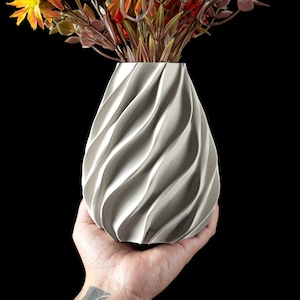 May include: A gray, teardrop-shaped vase with a textured, swirling design. The vase is filled with colorful faux flowers in shades of orange, yellow, and brown. The vase is held in a hand against a black background.
