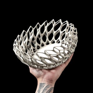 May include: A white ceramic bowl with a delicate, intricate, openwork design. The bowl is held in a hand, showcasing its delicate structure.