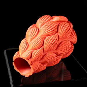 May include: A bright orange ceramic vase with a unique, textured design resembling overlapping leaves. The vase is lying on its side, revealing the open mouth.