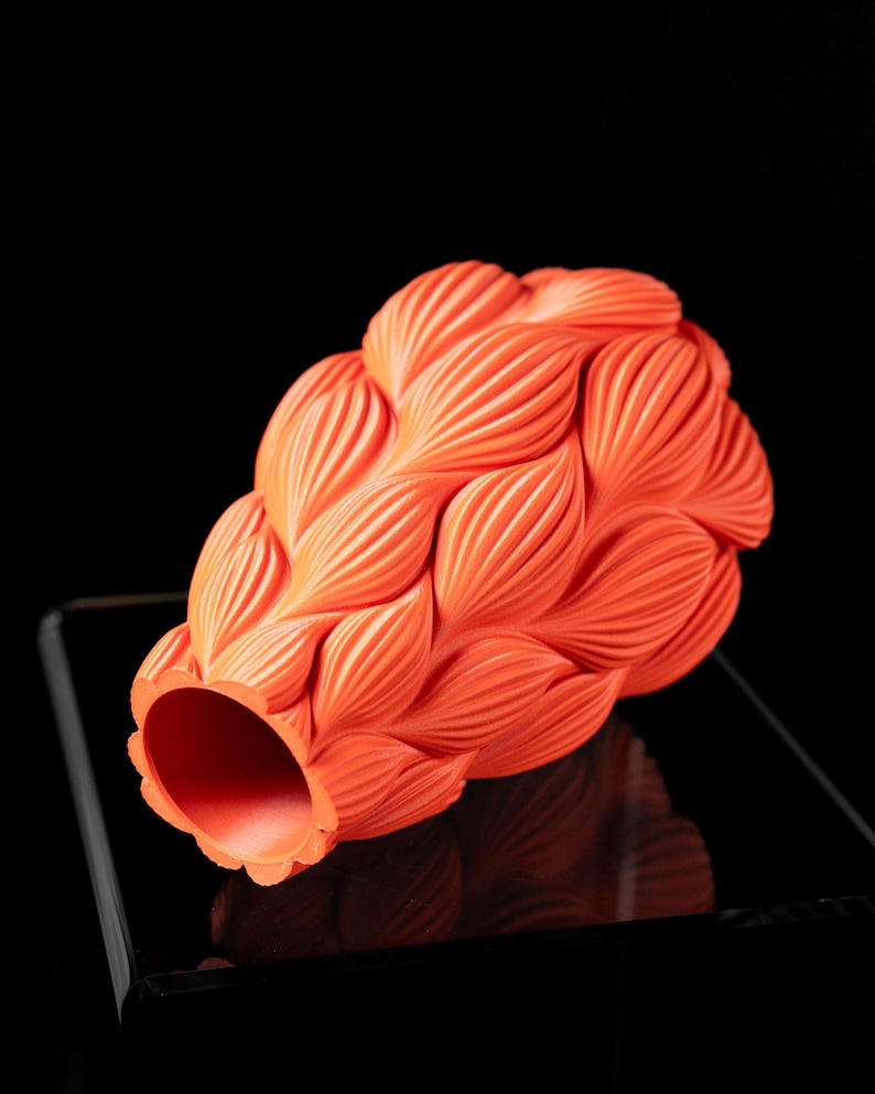 May include: A bright orange ceramic vase with a unique, textured design resembling overlapping leaves. The vase is lying on its side, revealing the open mouth.