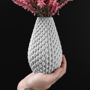 May include: A white vase with a distinctive scale-like design, held in a hand. The vase contains pink flowers, contrasting against the black backdrop. A decorative item for home interiors, with a unique texture.