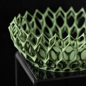 May include: A green, geometric, openwork bowl with a honeycomb pattern. The bowl is sitting on a black pedestal.