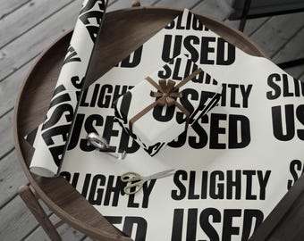 Könnte beinhalten: Cremefarbenes Geschenkpapier und ein verpacktes Geschenk, beide mit den Worten "SLIGHTLY USED" in fetten schwarzen Buchstaben bedruckt. Eine braune Schleife ziert das Geschenk. Eine Schere und Klebeband liegen ebenfalls auf dem Holztisch.