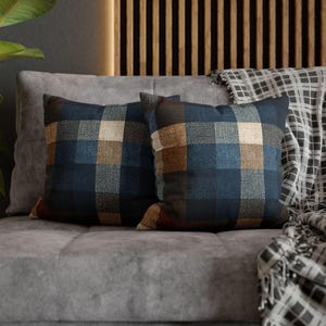 May include: Two square cushions with a blue, brown, and beige plaid design. The cushions are placed on a grey sofa, with a patterned throw draped over the back. The cushions add colour and texture to the neutral-toned living space.