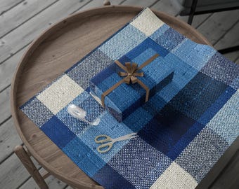 May include: A wrapped gift with a brown ribbon and bow sits on blue plaid wrapping paper. Scissors and tape are nearby on a wooden table. The wrapping paper has a textured appearance with shades of blue and cream.