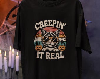 May include: Black t-shirt featuring a cat wearing sunglasses, set against a sunset design with pumpkins. The text "CREEPIN' IT REAL" is above and below the cat graphic. The shirt is held by a hand with long nails.