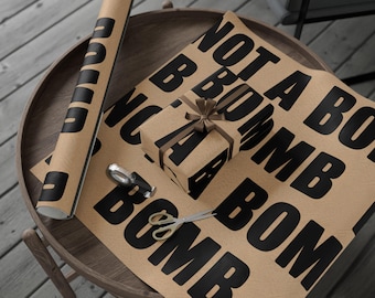 Könnte beinhalten: Braunes Geschenkpapier mit den Worten "BOMB" und "NOT A BOMB" in großen schwarzen Buchstaben. Eine Schere und ein Geschenk liegen auf einem Holztisch, mit einer Papierrolle.