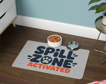 May include: A gray pet food mat with the words "SPILL ZONE ACTIVATED" in navy blue and orange. A white bowl filled with pet food and a silver bowl are on the mat. The mat is on a dark wood floor.