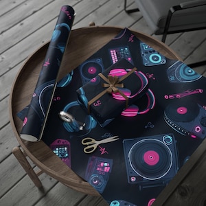 May include: Black wrapping paper with a neon music theme featuring headphones, turntables, and sound wave graphics. A wrapped gift with a brown ribbon sits on the paper, next to a roll of the same design. Gold scissors are also on the paper.