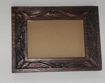 May include: A rectangular, dark brown wooden picture frame with ornate carved floral and scroll designs. The frame has a recessed center for a photo or artwork. The frame is approximately 10 inches wide and 8 inches tall.