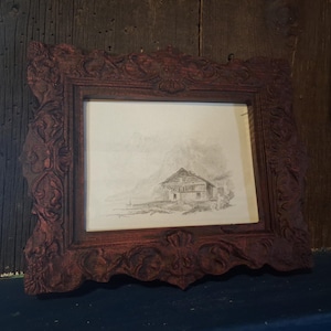 May include: A framed black and white print depicting a building in a landscape. The frame is a dark reddish-brown with detailed carvings. The print is centrally positioned and looks like an etching or drawing.