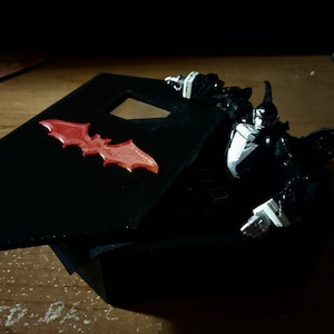 May include: A black coffin-shaped box with a red bat design on the lid. The box is open and a black and white action figure is inside.