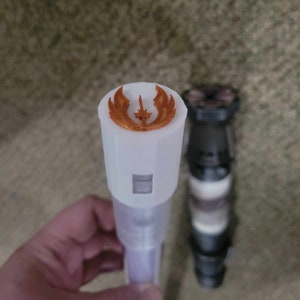 May include: A white lightsaber hilt with a copper-colored Jedi Order symbol on the top. The hilt is made of plastic and has a black and white design.