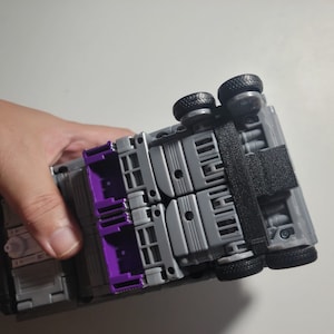 May include: A gray and purple toy truck with black wheels and a black strap. The toy is held in a hand and is upside down, revealing the underside of the vehicle.