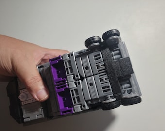 May include: A gray and purple toy truck with black wheels and a black strap. The toy is held in a hand and is upside down, revealing the underside of the vehicle.