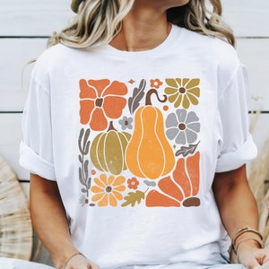 May include: White t-shirt with a graphic print of a square design featuring pumpkins, flowers, and leaves in orange, yellow, brown, and gray colors.