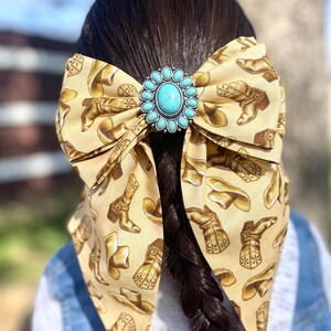 May include: A large hair bow made of tan fabric featuring a repeating pattern of brown cowboy boots and hats. The centre of the bow is adorned with a circular silver-toned concho featuring a central oval turquoise stone surrounded by small beads.