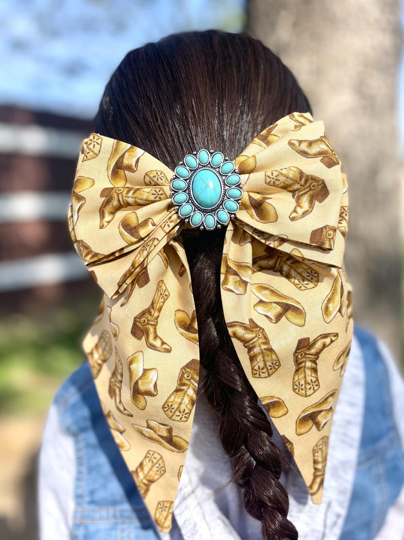 May include: A large hair bow made of tan fabric featuring a repeating pattern of brown cowboy boots and hats. The centre of the bow is adorned with a circular silver-toned concho featuring a central oval turquoise stone surrounded by small beads.