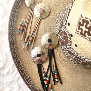 May include: Two pairs of dangle earrings featuring scalloped silver-tone metal concho discs. One pair has tan suede fringe with wooden, red, and turquoise-colored beads. The other pair has black suede fringe with similar bead accents.