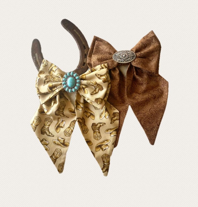 May include: Two fabric bows attached to a dark metal horseshoe. One bow is cream-coloured with a brown cowboy boot and hat print, featuring a central turquoise stone. The other bow is solid brown with a tonal paisley pattern and a silver oval concho.