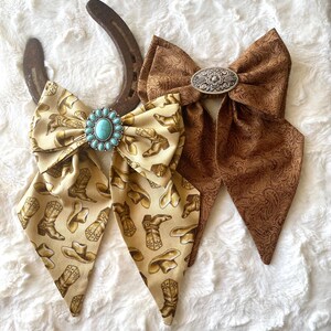 May include: Two fabric bows with long tails. One bow is cream-coloured with a brown cowboy boot and hat print, featuring a turquoise stone concho. The other bow is brown with a tonal paisley pattern and a silver-toned oval metal concho.