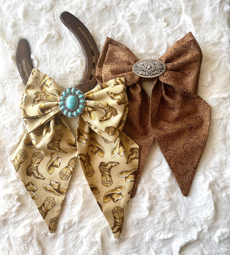 May include: Two fabric bows with long tails. One bow is cream-coloured with a brown cowboy boot and hat print, featuring a turquoise stone concho. The other bow is brown with a tonal paisley pattern and a silver-toned oval metal concho.