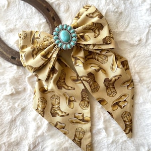 May include: A beige fabric bow featuring a repeating pattern of brown cowboy boots and hats. The centre of the bow is adorned with an oval turquoise stone set in a silver-toned metal frame with small decorative beads, positioned over a horseshoe.