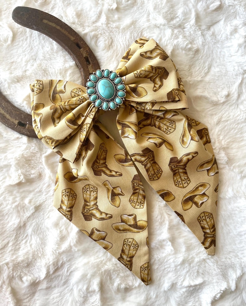 May include: A beige fabric bow featuring a repeating pattern of brown cowboy boots and hats. The centre of the bow is adorned with an oval turquoise stone set in a silver-toned metal frame with small decorative beads, positioned over a horseshoe.