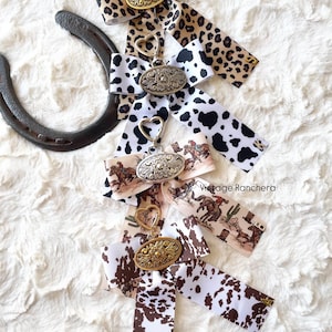 May include: Keychains with decorative ribbons in various patterns, including leopard print, cow print, and a cowboy scene. Each keychain features a metal oval pendant and a heart-shaped clasp. A horseshoe is in the background.