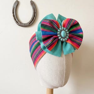 May include: A colorful baby headband with a large bow. The bow features a vibrant striped pattern in shades of pink, orange, blue, and green, with a turquoise center. The headband is displayed on a mannequin head.