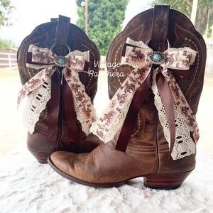 May include: A pair of brown leather cowboy boots with decorative bows. The bows feature a western-themed print with a turquoise center and lace trim. The boots are set on a white, textured surface.