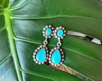 May include: A pair of turquoise dangle earrings with silver settings. The turquoise stones are a bright blue colour and are set in a teardrop shape. The earrings are surrounded by a silver halo with a starburst design.