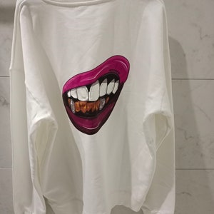 Pu&ograve; includere: Felpa bianca con una grafica di bocca di cartone animato rosa e oro sul retro.