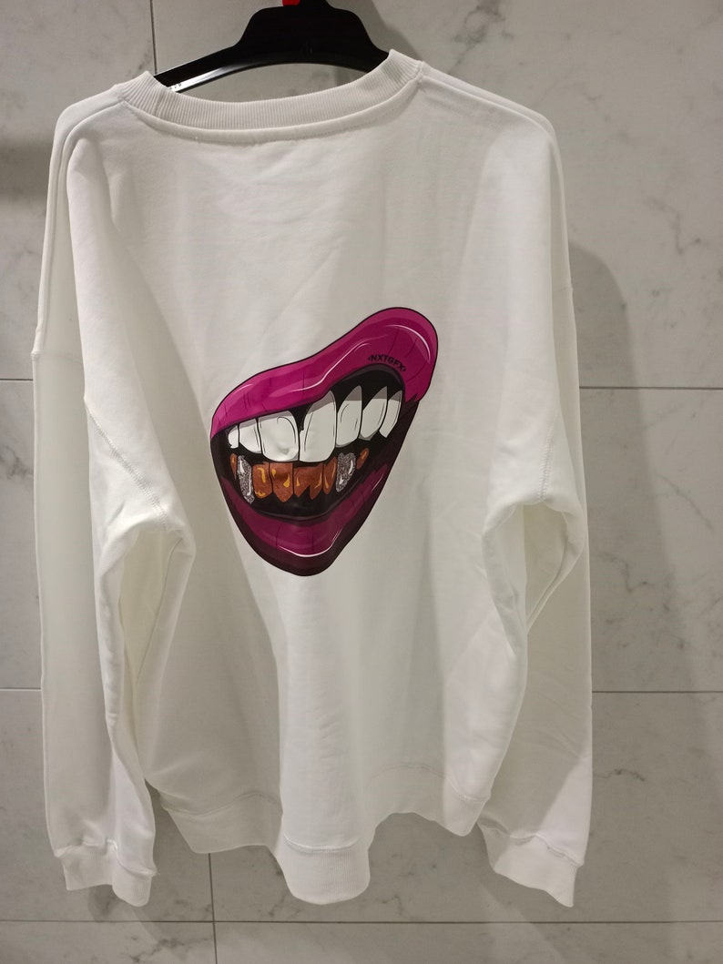 Pu&ograve; includere: Felpa bianca con una grafica di bocca di cartone animato rosa e oro sul retro.