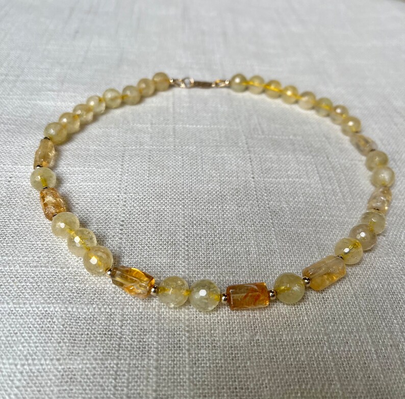 May include: A gold necklace made of light yellow and amber colored beads. The necklace is made of round beads and rectangular beads.