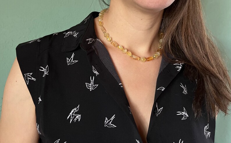 May include: A close-up of a woman wearing a black sleeveless shirt with a white bird pattern and a yellow amber necklace.