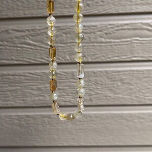 May include: A gold and clear crystal beaded necklace with a gold clasp. The necklace is made of round and rectangular beads.