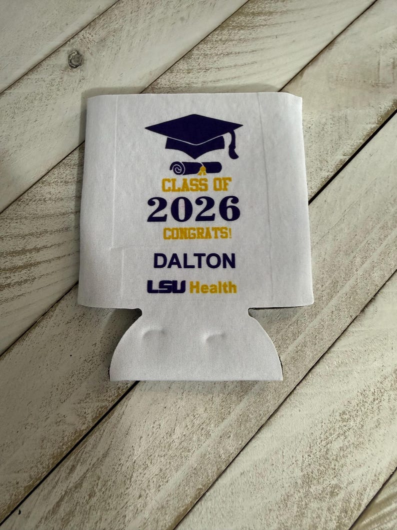 May include: White graduation can cooler with a navy blue mortarboard and scroll design. The text reads "CLASS OF 2026 CONGRATS! DALTON LSU Health" in yellow and navy blue. The cooler has a cutout for a can or bottle.
