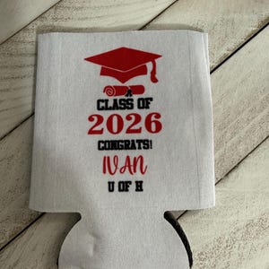 May include: White fabric can cooler with a red graduation cap and scroll design. Text includes "Class of 2026", "Congrats!", the name "Ivan", and "U of H". The can cooler has a black bottom.