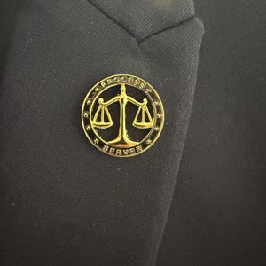 May include: A gold-coloured lapel pin featuring the words "PROCESS SERVER" around a scales of justice symbol. The pin is circular with a star design around the edge. It is attached to a black blazer.