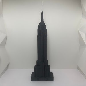 May include: A black miniature replica of the Empire State Building. The model is a detailed representation of the iconic skyscraper, featuring its distinct Art Deco design and spire. The building is set against a white background.