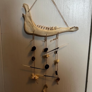 May include: A decorative wall hanging featuring a bleached animal jawbone suspended by twine. Below the jawbone, various natural elements like porcupine quills, small bones, and dark beads are strung, creating a rustic, earthy aesthetic.