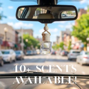 May include: A small, clear glass car air freshener bottle with a light-coloured wooden cap and a braided hanging cord, suspended from a rearview mirror. The text 40+ SCENTS AVAILABLE! is overlaid in white against the blurred street background.