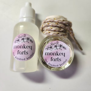 May include: Two clear glass bottles with a pink and white label that says "monkey farts the kindred krafter". The smaller bottle has a gold cord tied around the top.
