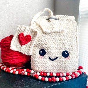 May include: White crocheted backpack with a cute cartoon face, red yarn, and a red heart-shaped crocheted patch. The backpack has a white and red beaded garland around it.