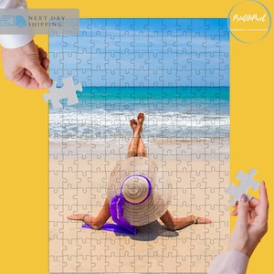 May include: A woman wearing a large straw hat with a purple ribbon lies on a sandy beach with her feet in the air. The ocean is in the background.