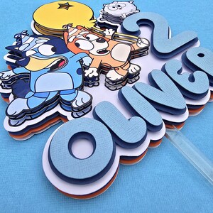 May include: A blue, orange, and white cake topper with the name "Oliver" and the number "2" on it. The topper features cartoon characters from the show "Bluey".