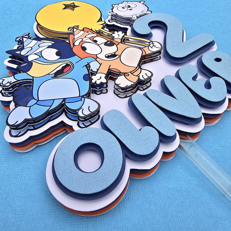 May include: A blue, orange, and white cake topper with the name "Oliver" and the number "2" on it. The topper features cartoon characters from the show "Bluey".