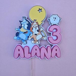 May include: A pink and white cake topper with the name "Alana" and the number "3" in pink. There are two cartoon dogs holding a yellow balloon with a star on it. There is also a white cartoon dog with a pink nose.