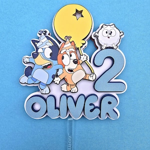 May include: Blue, orange, and white cartoon characters on a blue and white cake topper with the number 2 and the name Oliver.